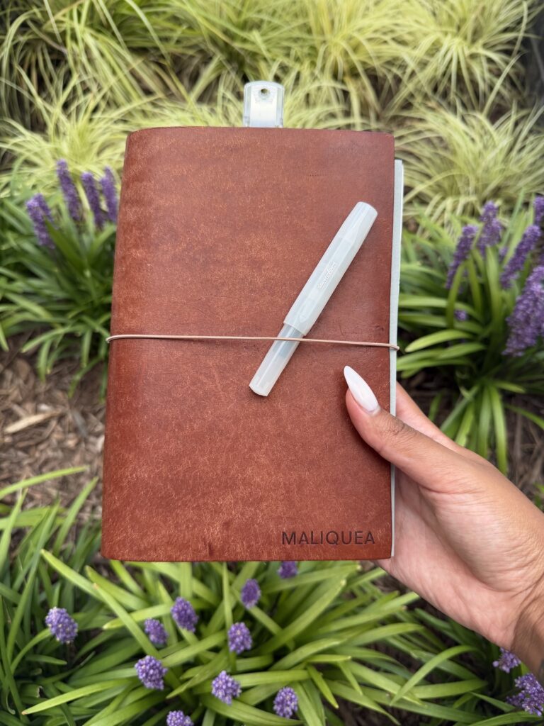 My personalized tan leather journal cover, embossed with my name, held up against a garden of purple flowers.